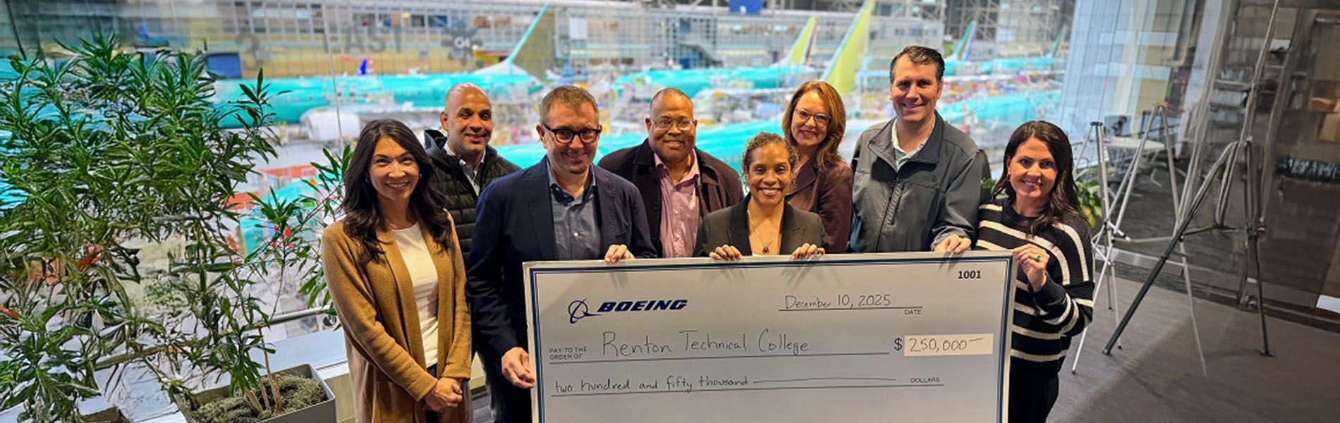 Group of eight individuals presenting a large check from The Boeing Company to Renton Technical College for $250,000, with an indoor construction site in the background.