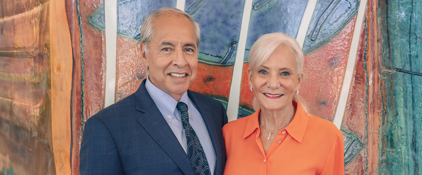 Two adults smiling and standing together in front of a colorful abstract painting. One adult is wearing a navy blue suit and tie, and the other is dressed in a bright orange shirt.