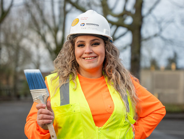 A construction student wearing a hardhat and a neon vest holding a paint brush