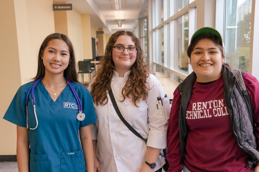 Three RTC Running Start students smiling and wearing scrubs, a chef's coat, and RTC gear.